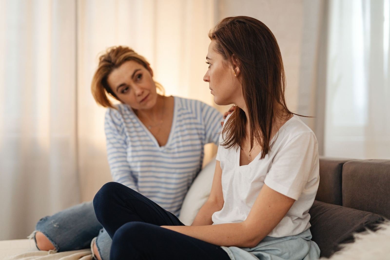 One woman sits on a bed with another. She puts a caring hand on her friend's shoulder.