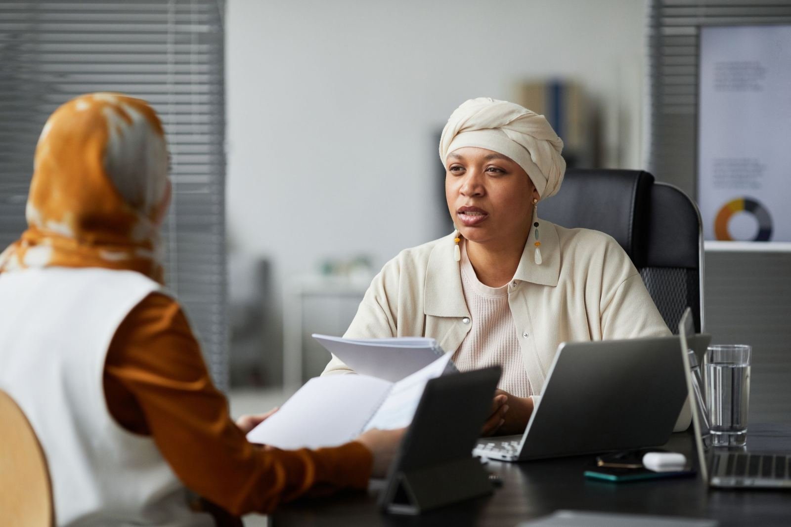 In a bright office, two women sit at a desk with papers and a laptop.