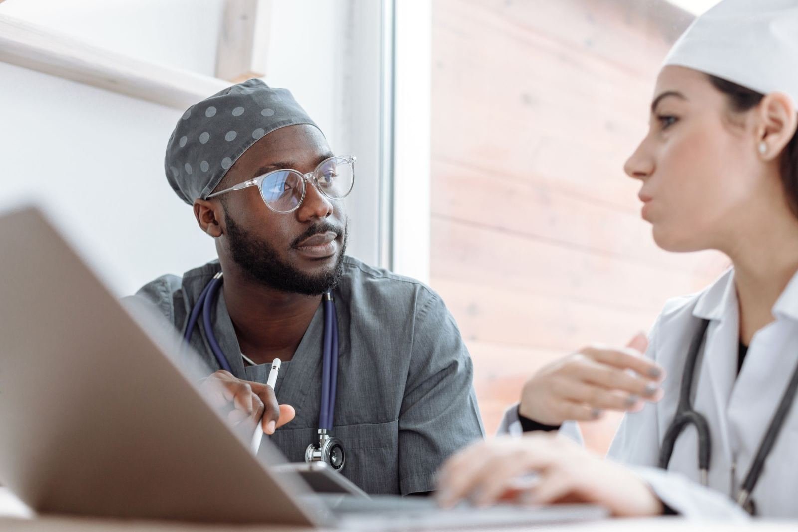Two medical professionals in scrubs and a lab coat discuss information on a laptop