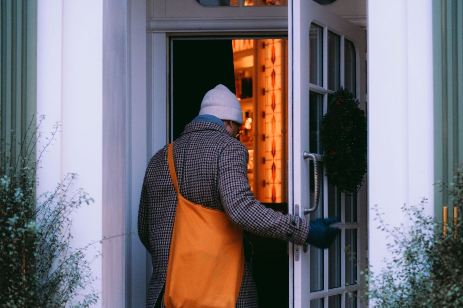 A person in a patterned coat and orange bag opens the door to a brightly lit home