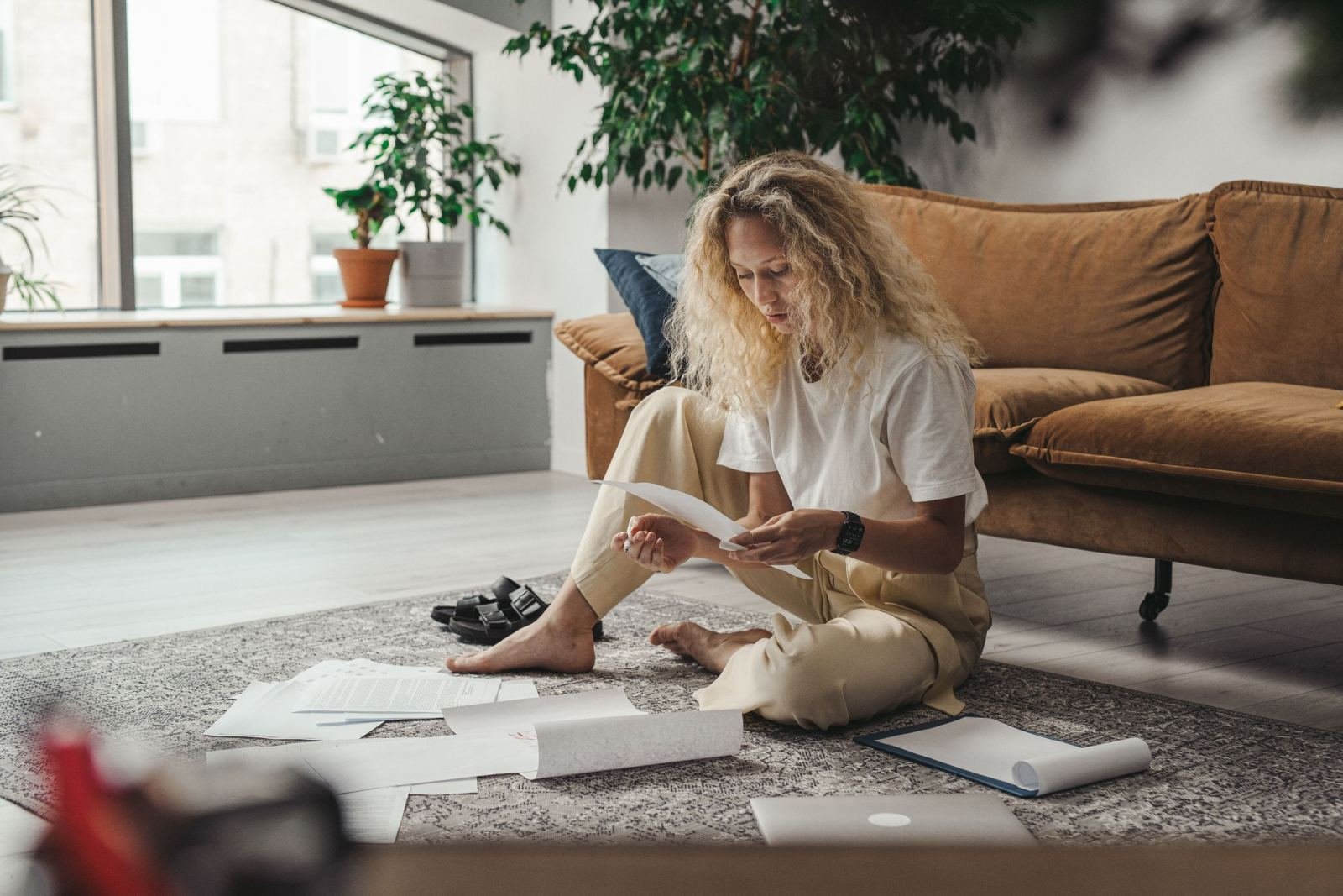 A woman sits on a rug reading papers with a laptop and documents around her