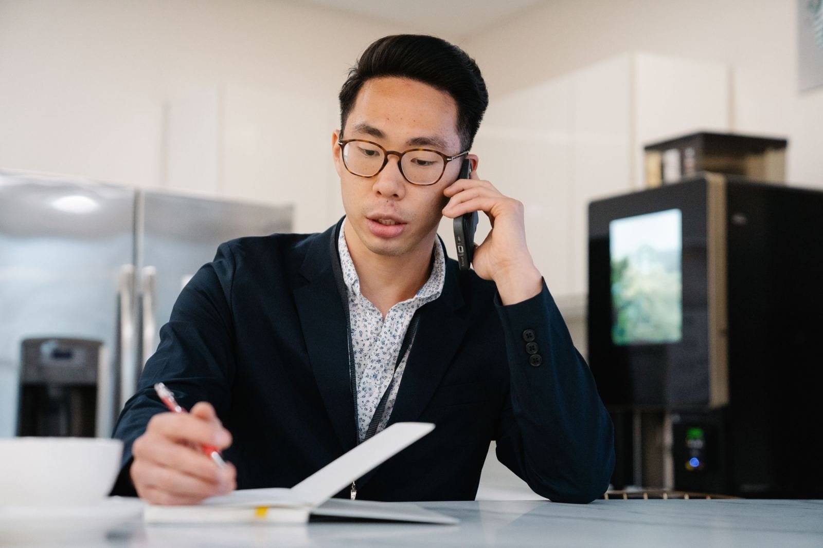 A man in a blazer speaks on the phone while writing in a notebook