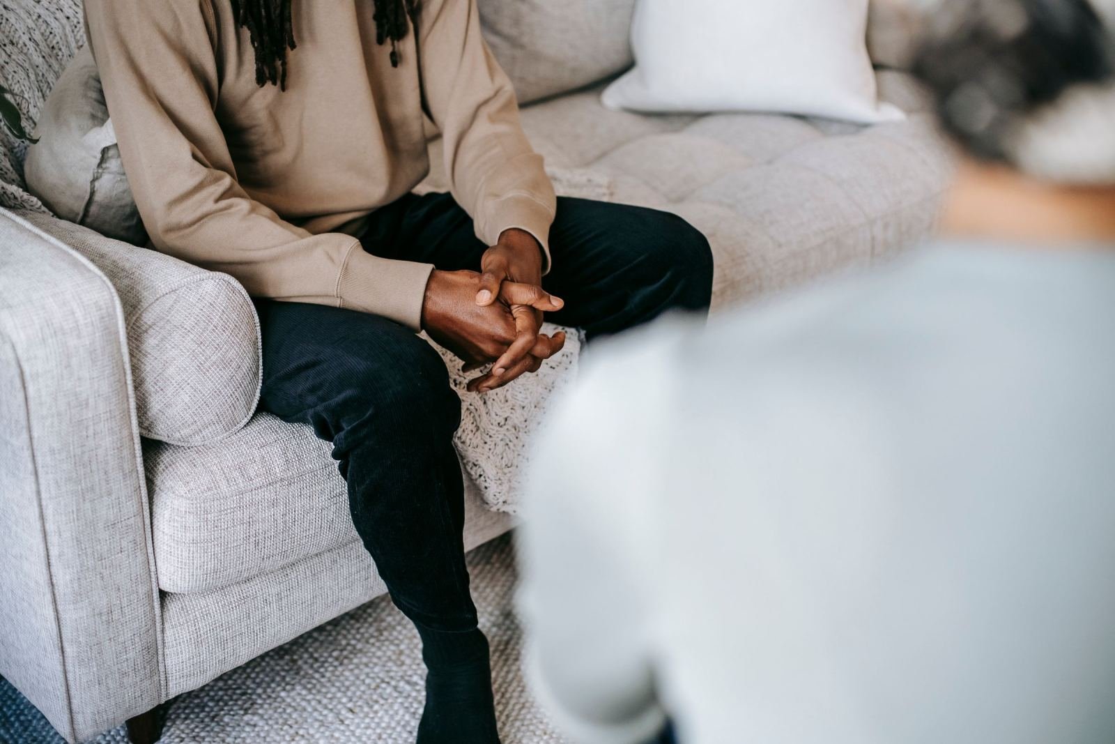 A person sits on a sofa with clasped hands while talking to someone