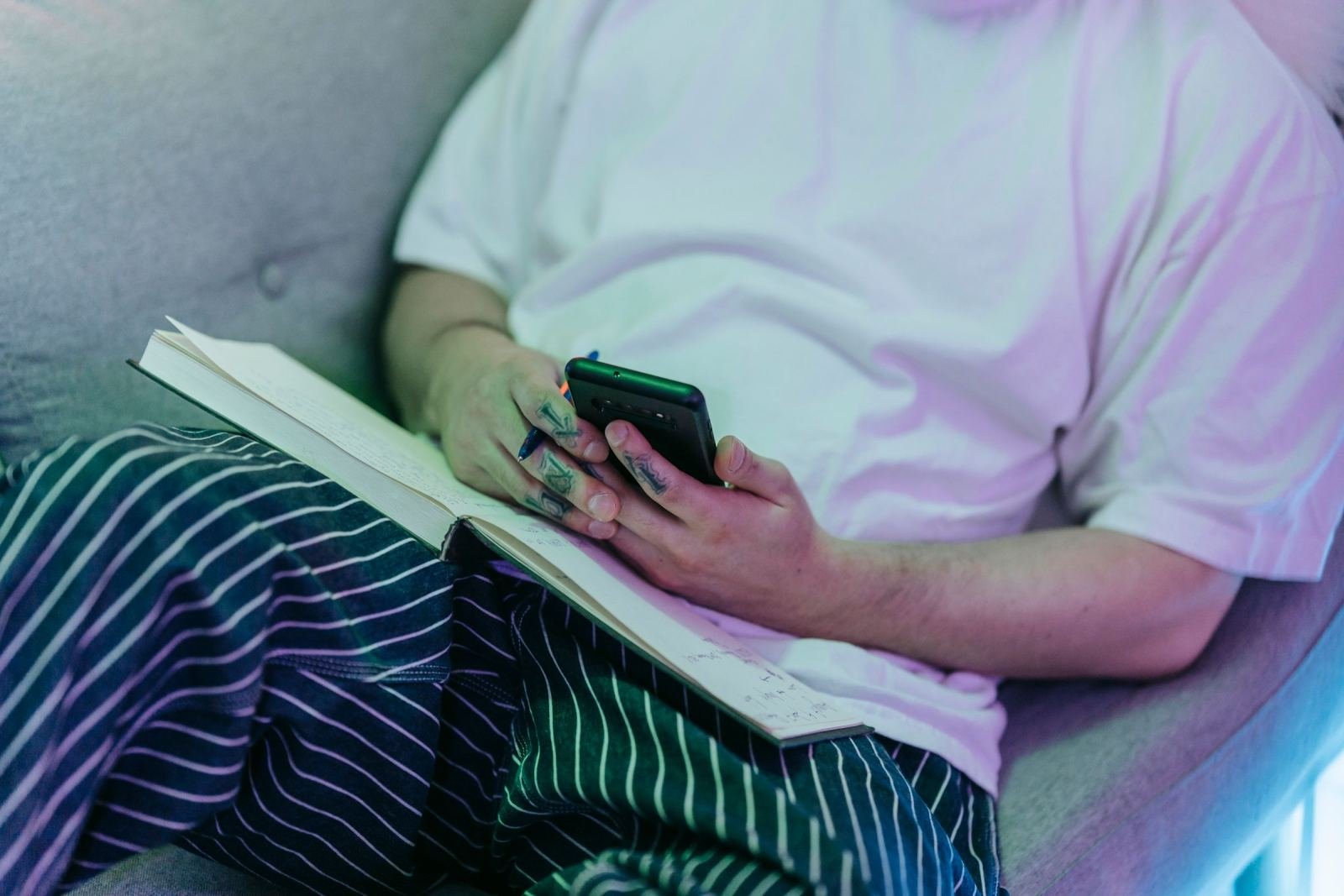 A person sits on a sofa holding a smartphone. They also hold a pen with a notebook on their lap.