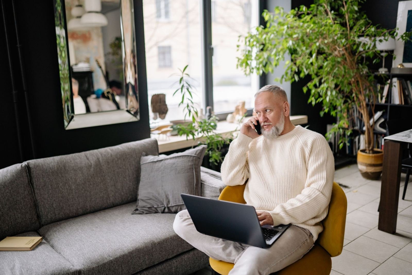 A man wearing a white sweater is on the phone and using a laptop in a living room