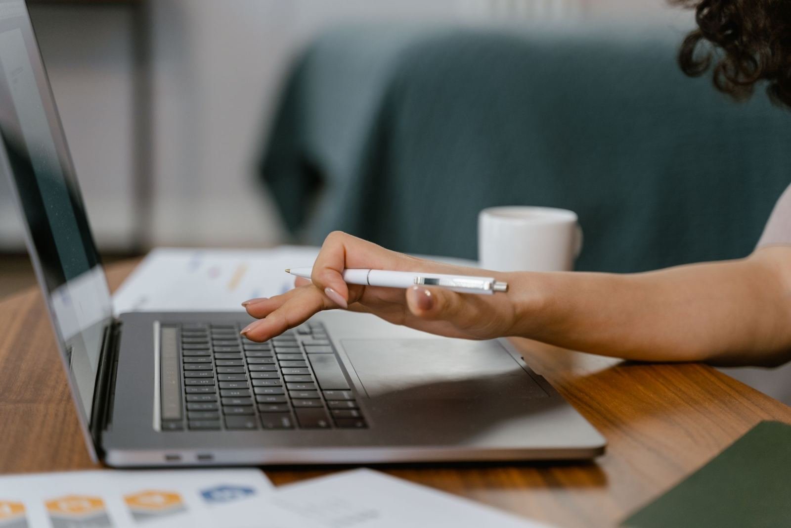 A person using a laptop and pen to review documents on a wooden table