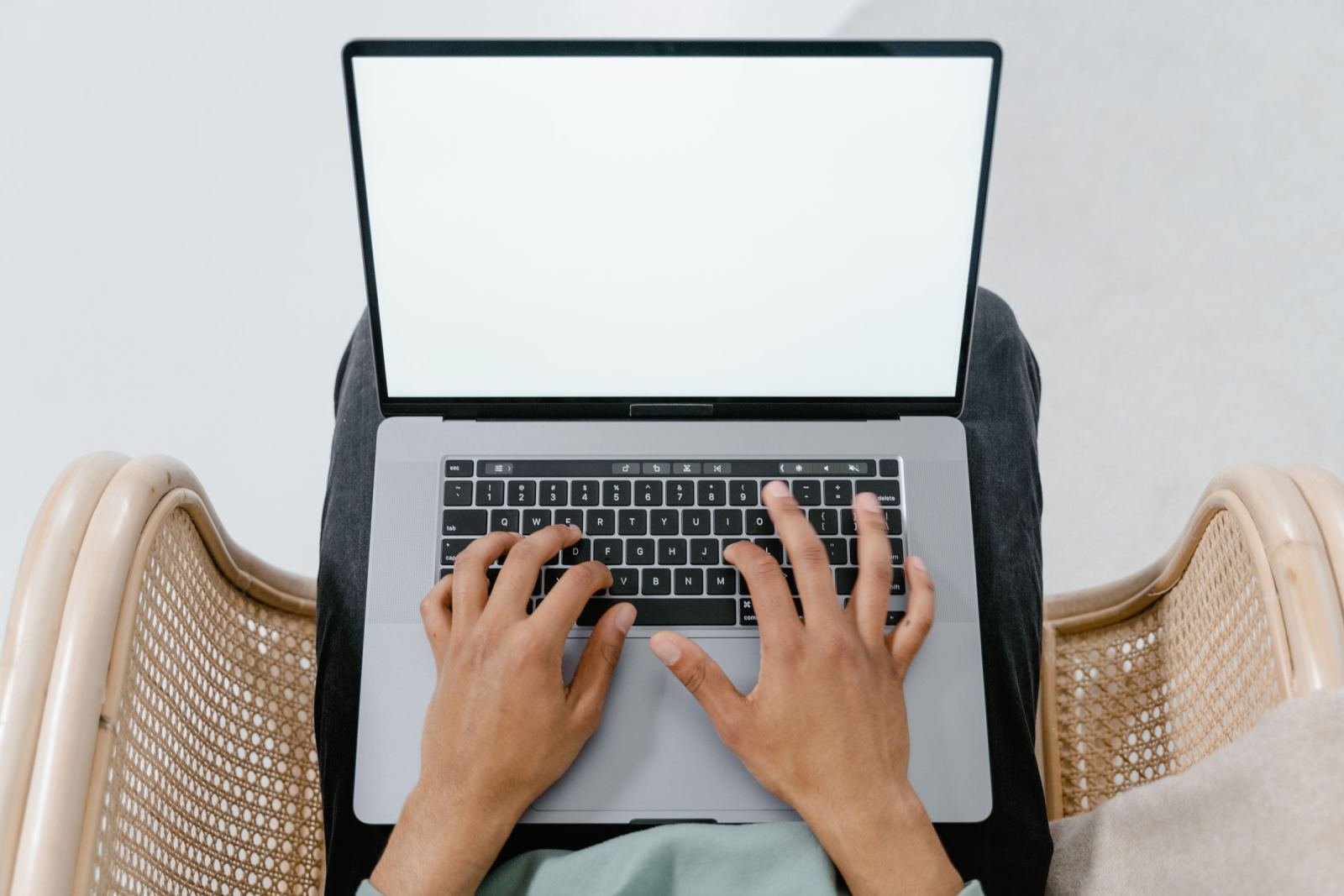 A person typing on a laptop with a blank white screen while sitting in a wicker chair