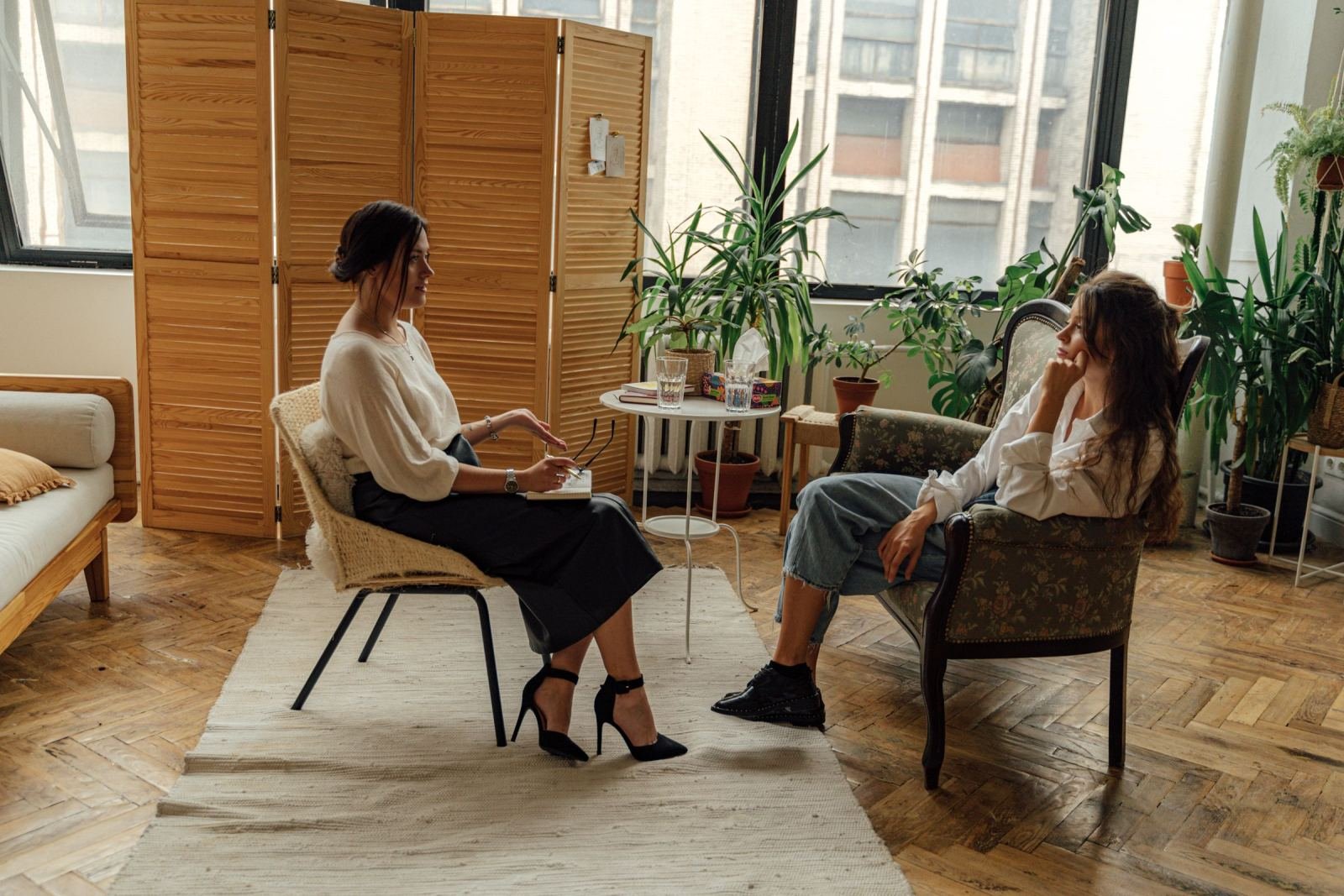 Two women sit in chairs facing each other in a bright room with plants