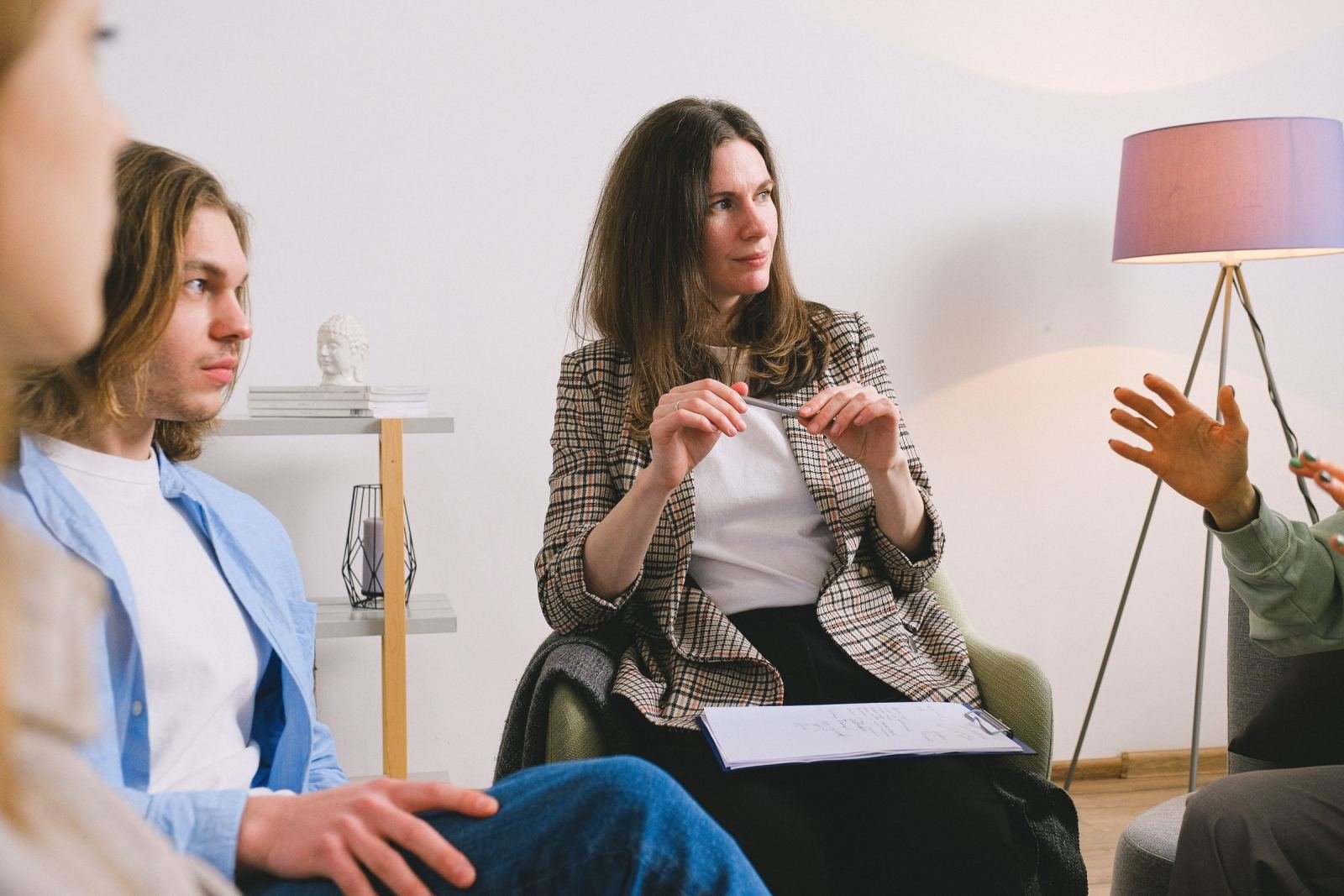 A woman in a plaid blazer sits in an office with two others, speaking and gesturing.
