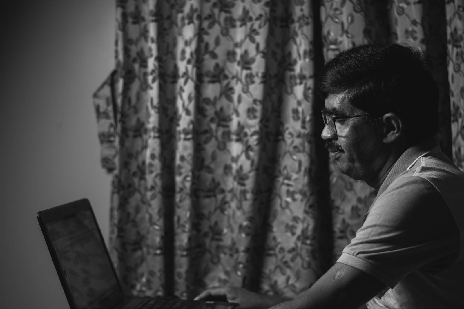 A man wearing glasses looks at his laptop screen near a patterned curtain
