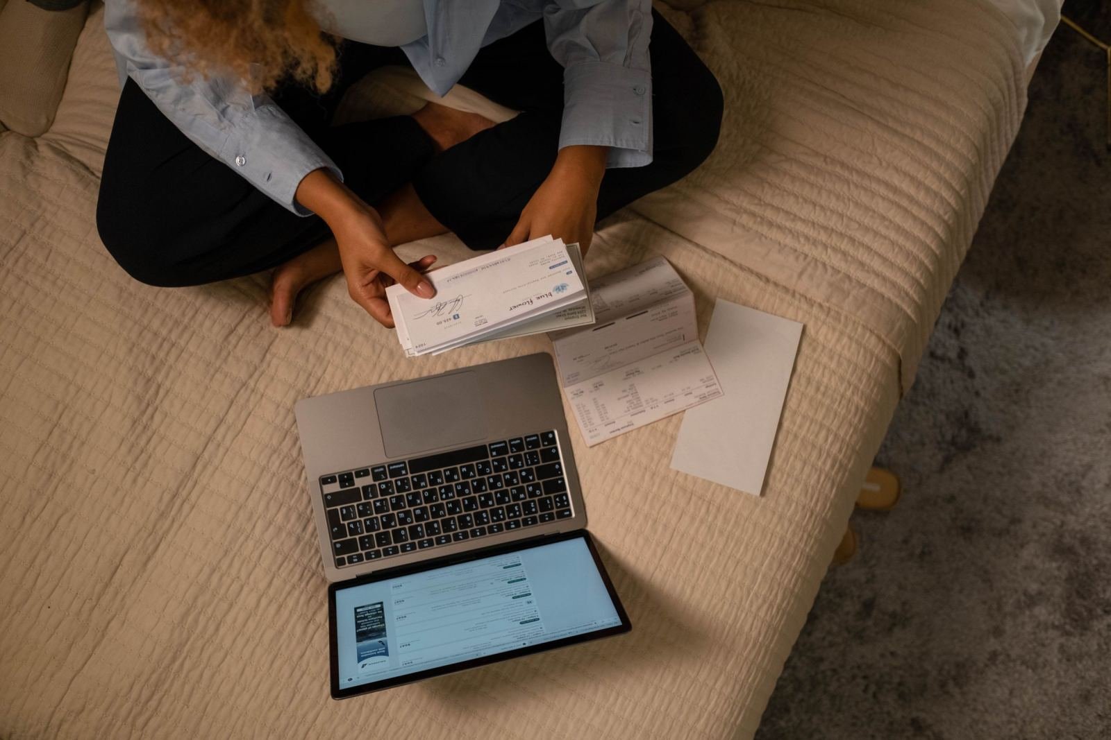 A person sits on a bed with a laptop and a stack of envelopes and papers