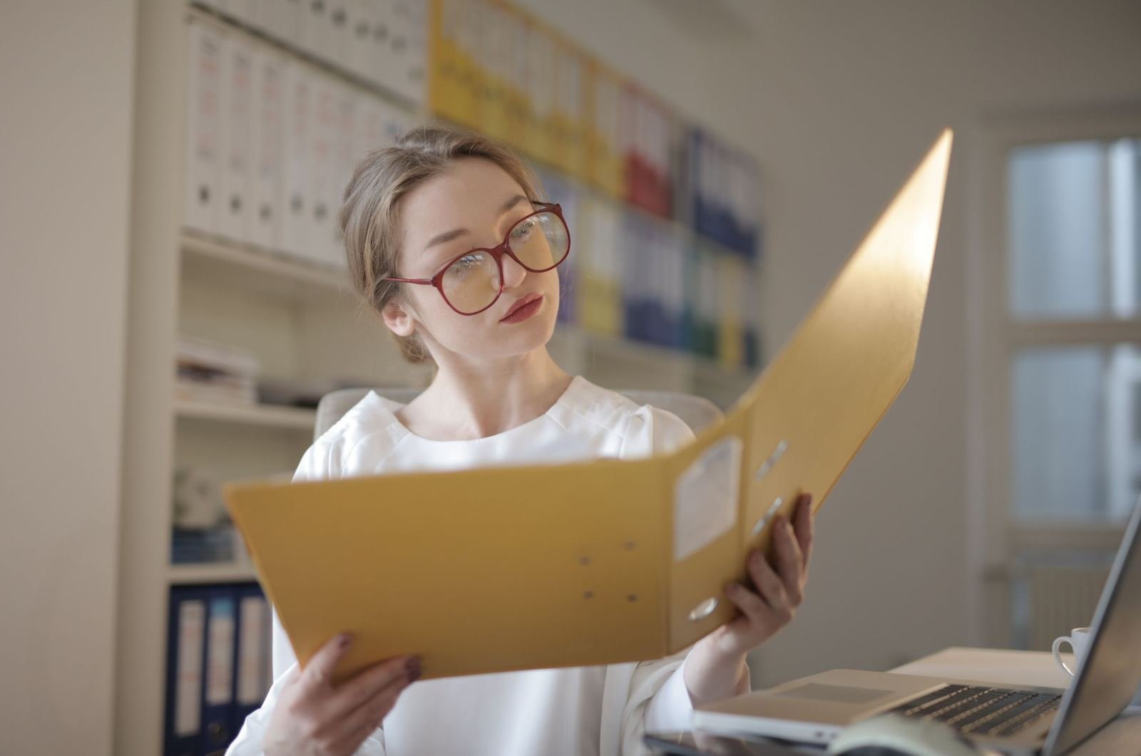 A woman with red glasses reviews a yellow binder at her desk