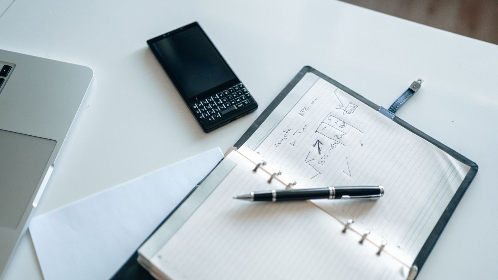 A laptop, smartphone, and open binder with handwritten notes on a white desk