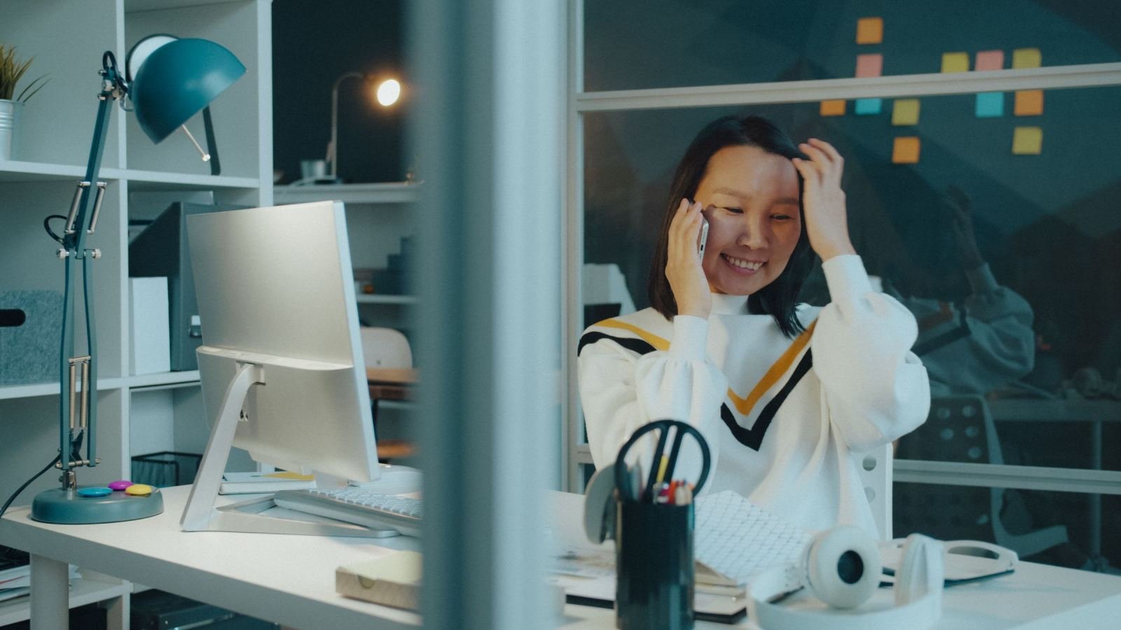 A happy woman sits at her work desk and chats on a cell phone.