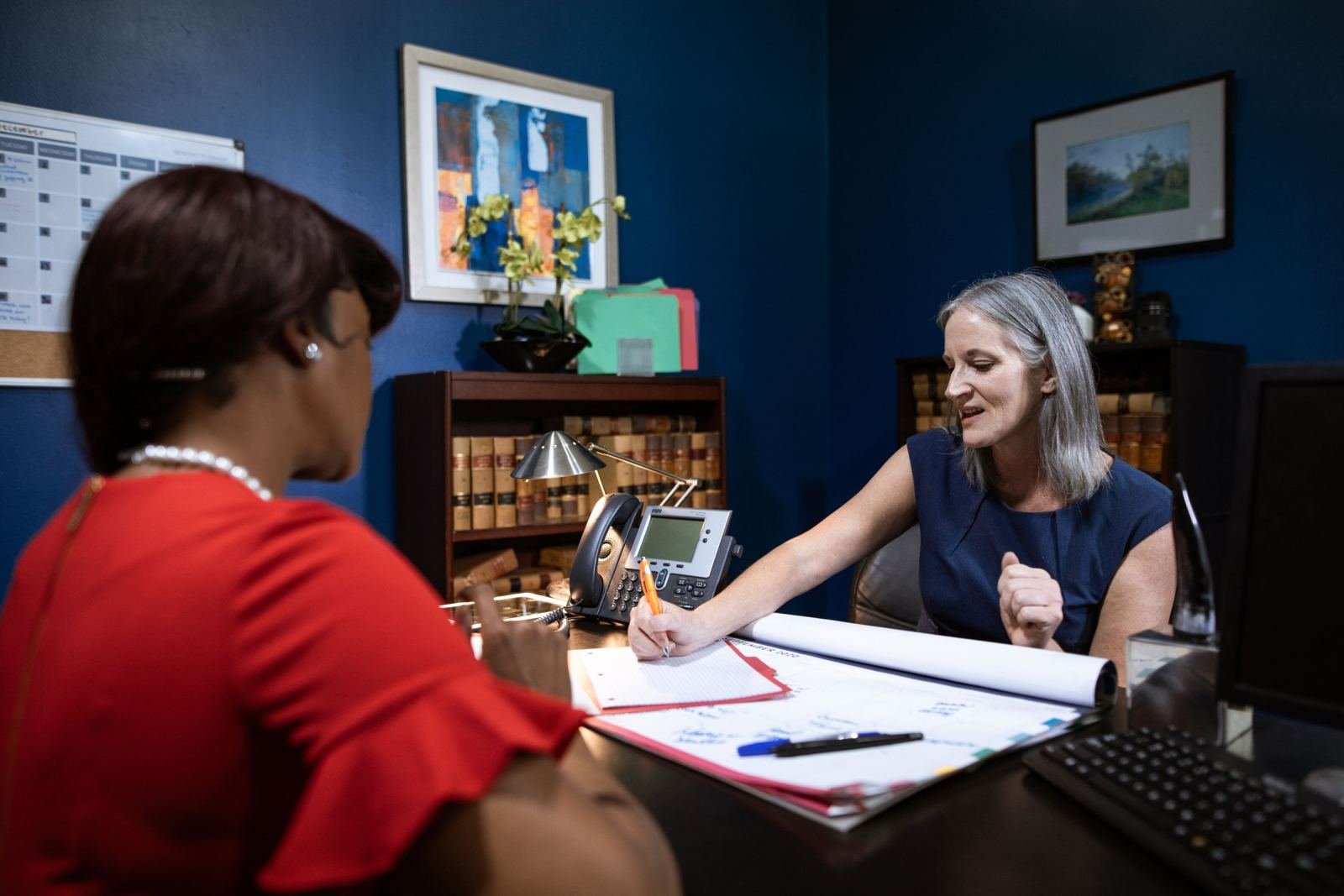 A woman in a red top talks with a counselor in a blue top over a desk.