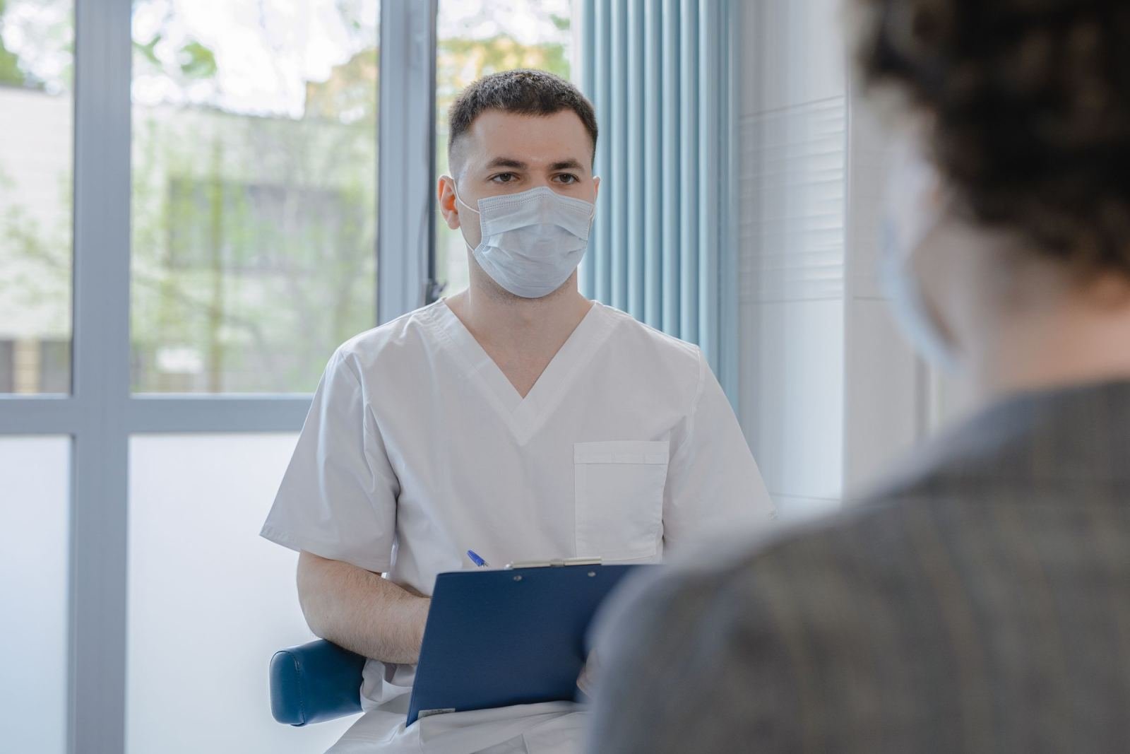 A medical professional in a mask and scrubs holds a clipboard while speaking with a patient