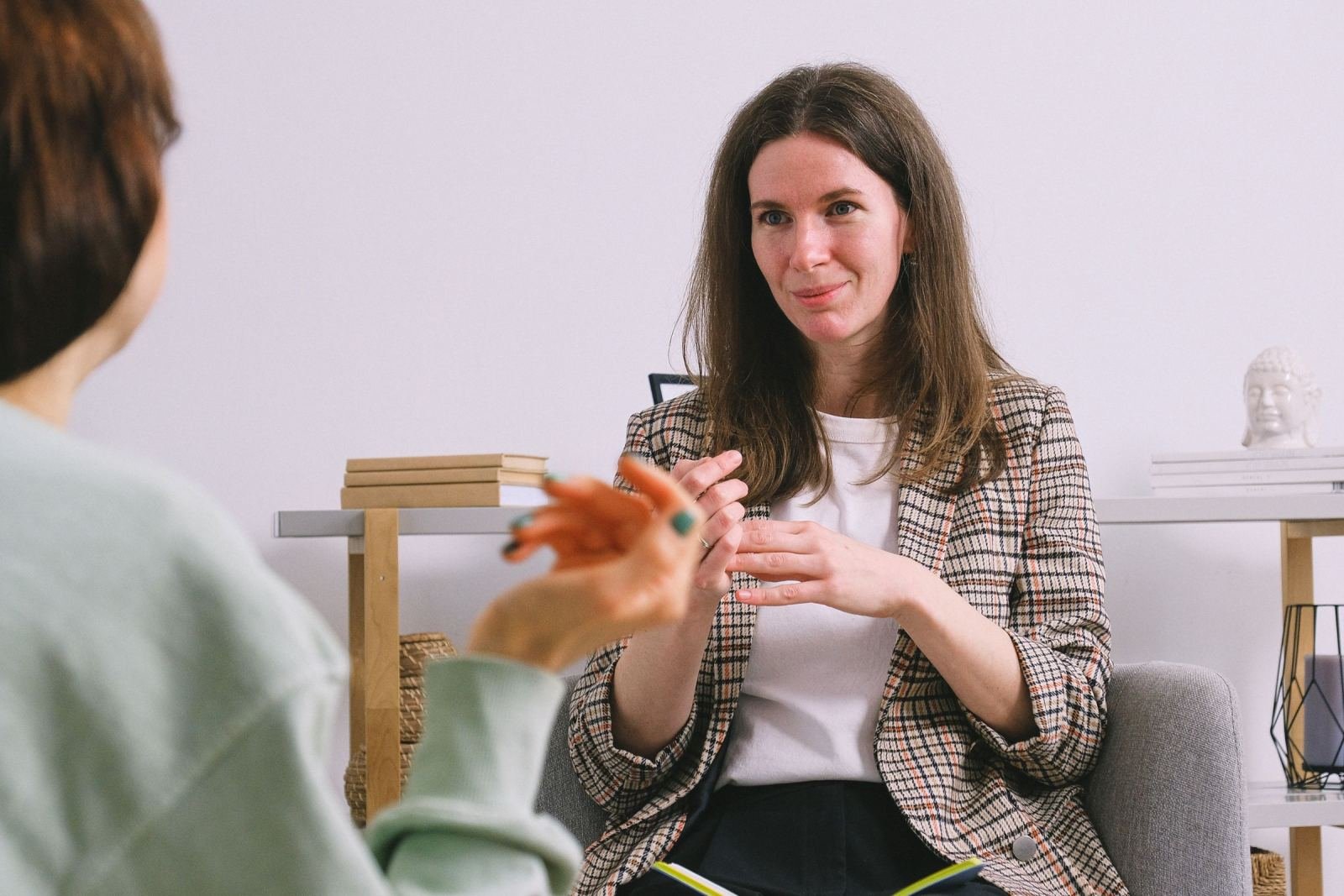 A woman in a plaid blazer gestures while speaking to a person seen from behind