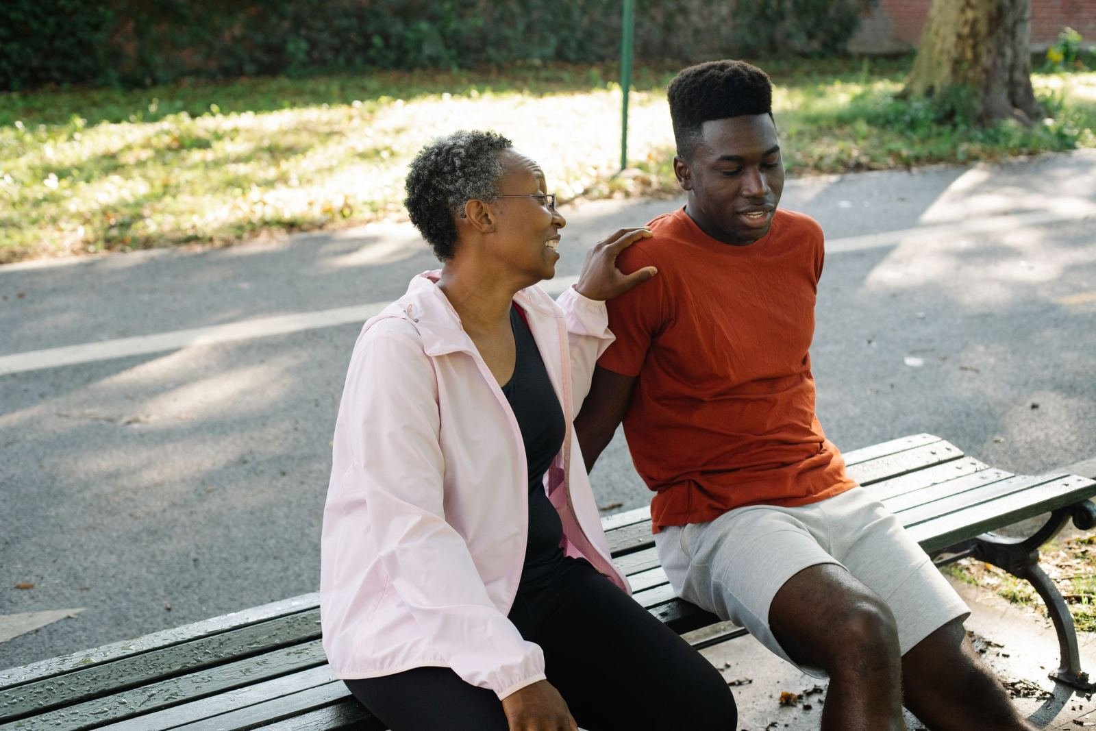 A young man sits on a park bench while an older woman rests her hand on his shoulder.