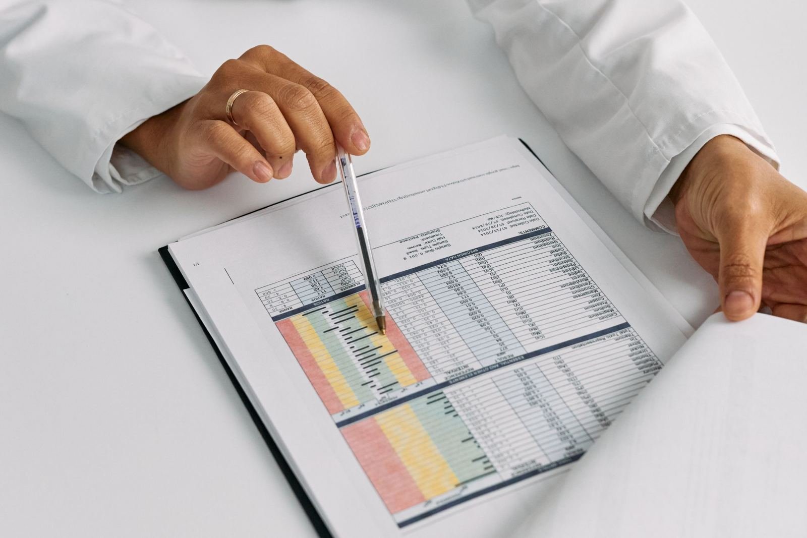 A person in a white coat points to a data table in a document with a pen