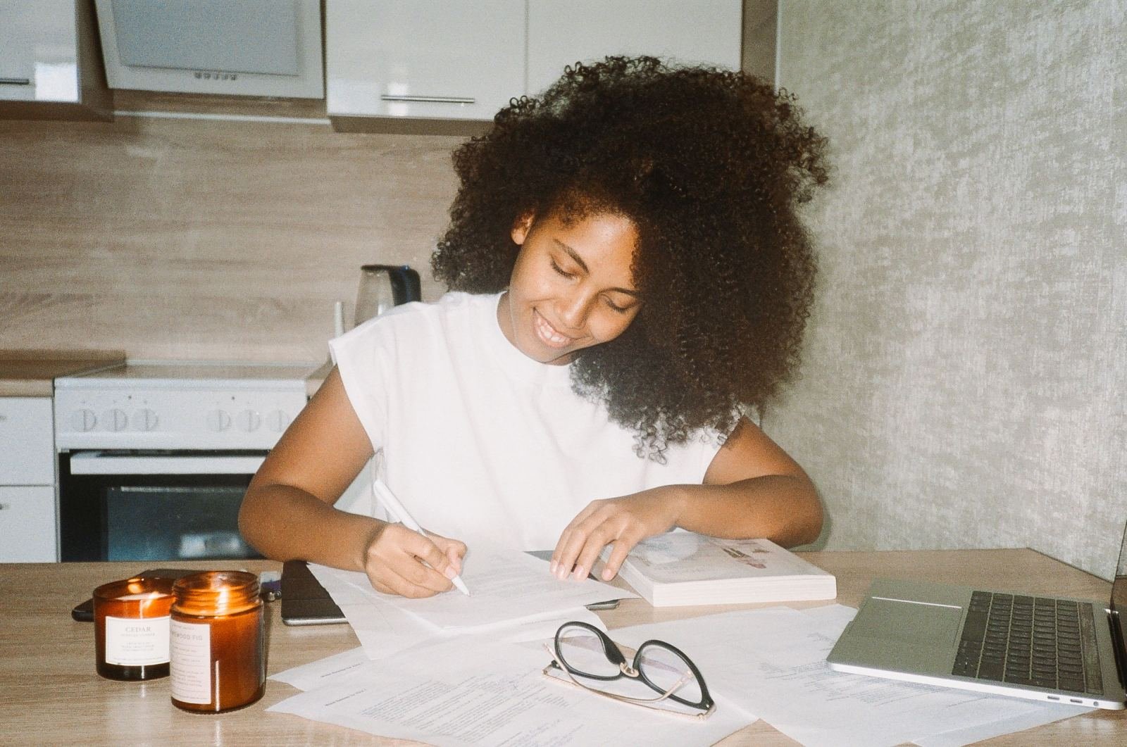 A woman writes in a notebook at a kitchen table with a laptop and candles