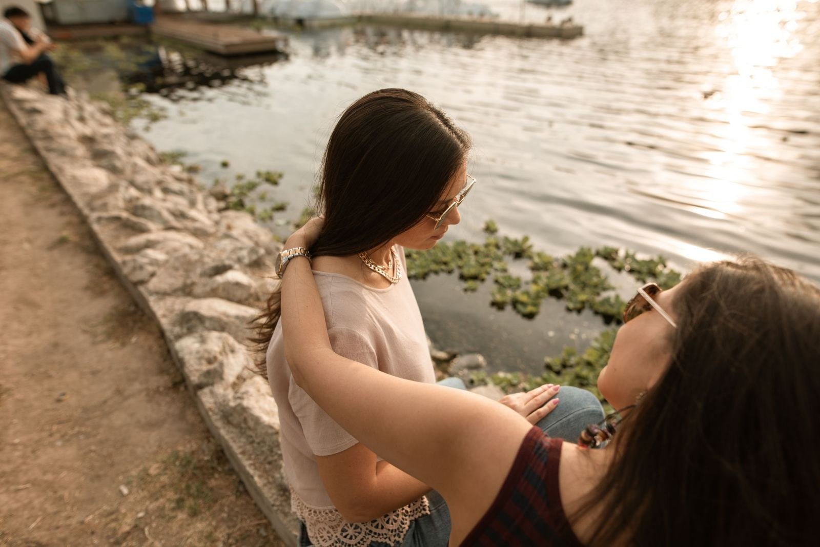 One woman puts her arm around another woman's shoulder while sitting by a pond