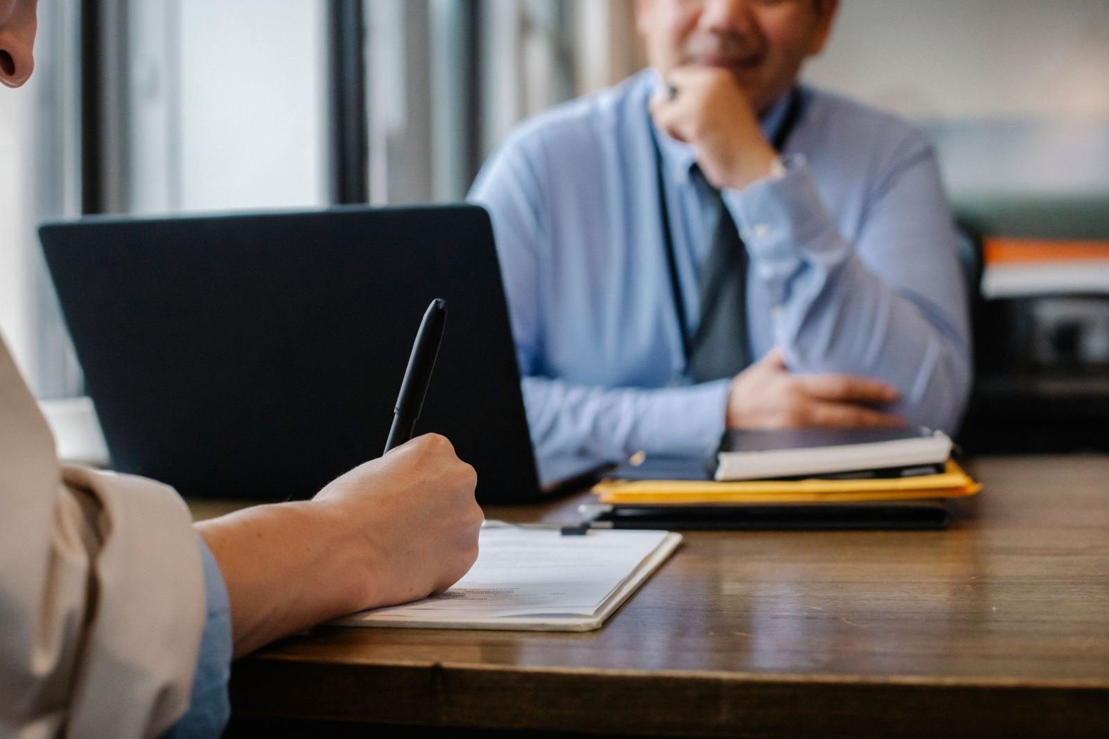 A person writes in a notebook while talking with a man in a blue shirt and tie