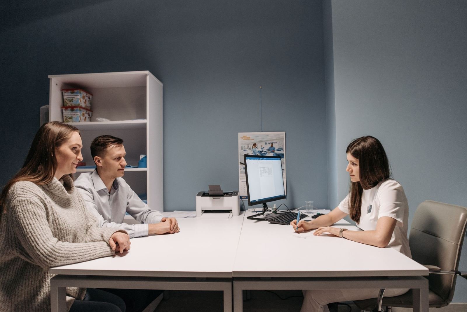 A medical professional in white scrubs speaks with a couple at a desk