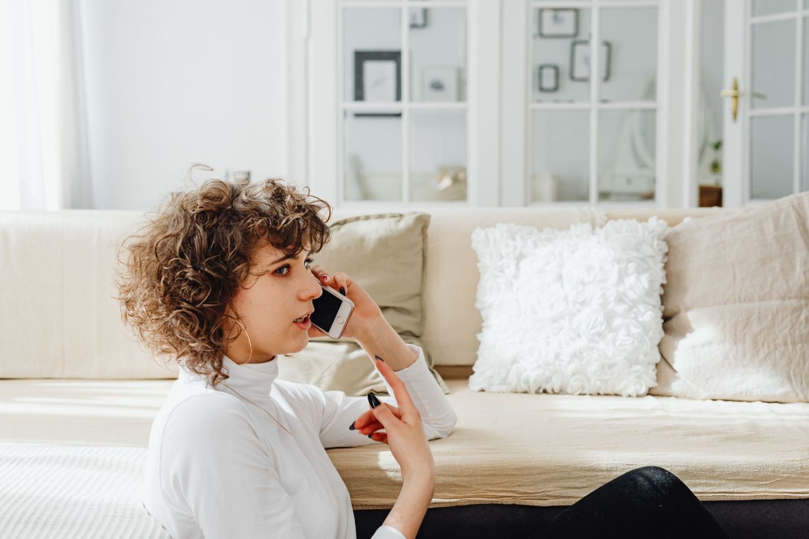 A woman with curly hair sits on a sofa while talking on her smartphone.