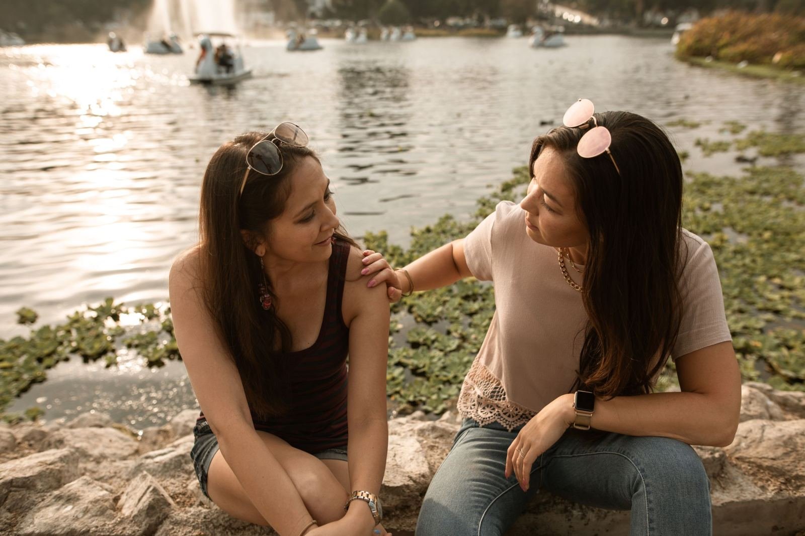 Two women talk on a stone ledge near a lake. One rests her hand on the other's shoulder.