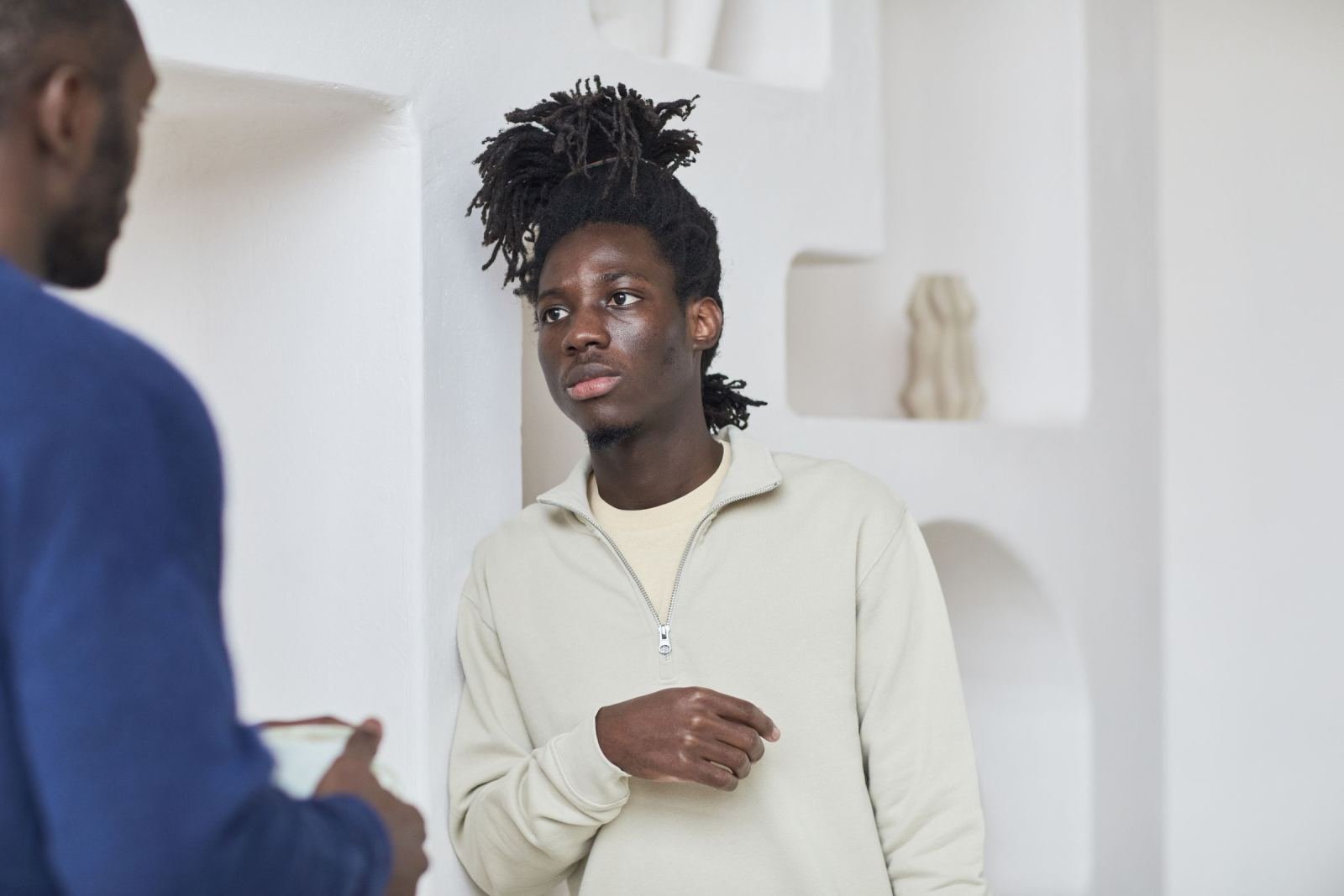 A young man with dreadlocks leans against a white wall while talking to another man
