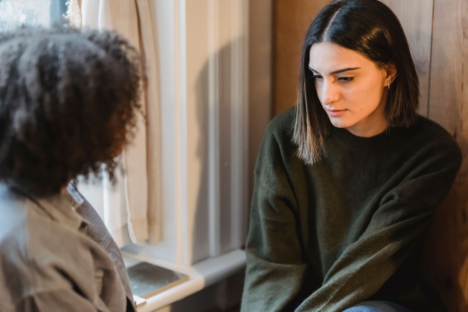 A woman in a green sweater looks down while talking to another person