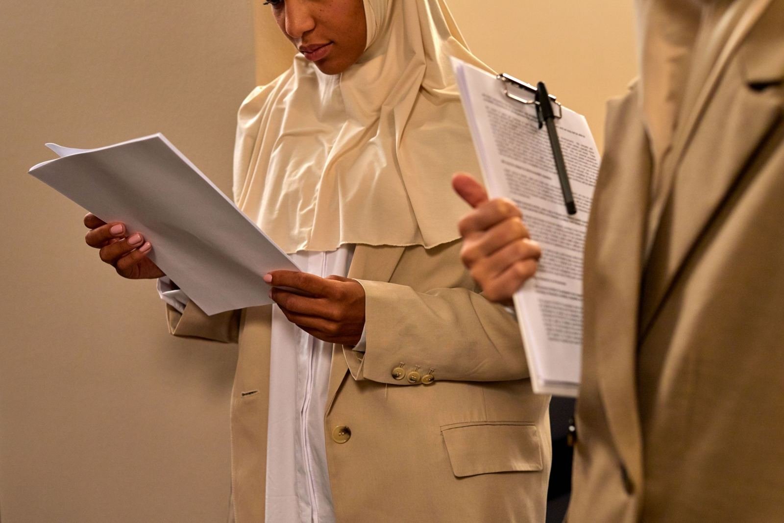 A woman in a hijab and tan blazer reads a document while standing next to a colleague with a clipboard