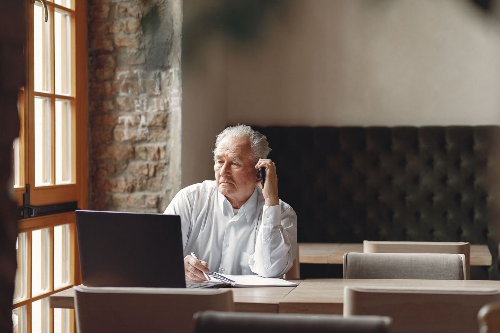 A senior man makes a phone call while sitting at a desk with a laptop and a notepad
