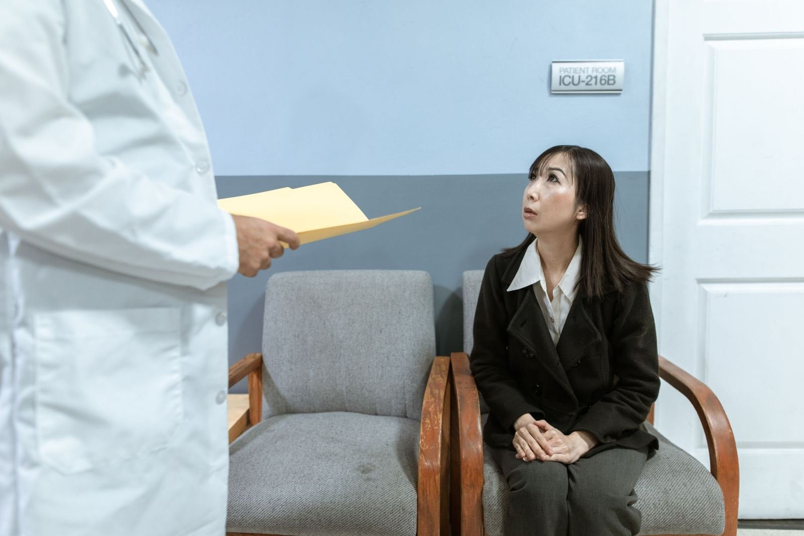 A doctor in a white coat speaks with a woman sitting in a grey chair