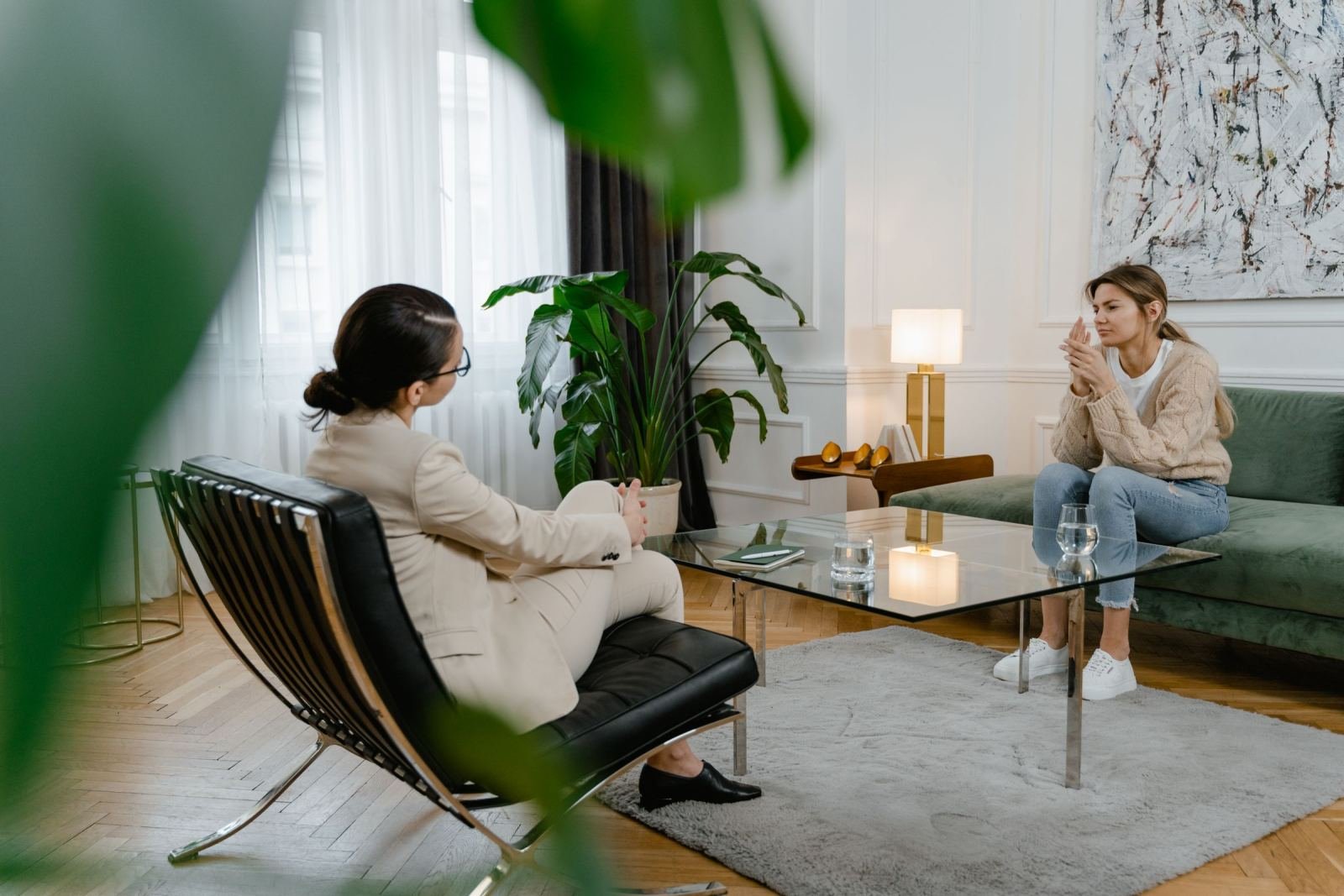 A woman in a suit talks with a client sitting on a green sofa in a bright office