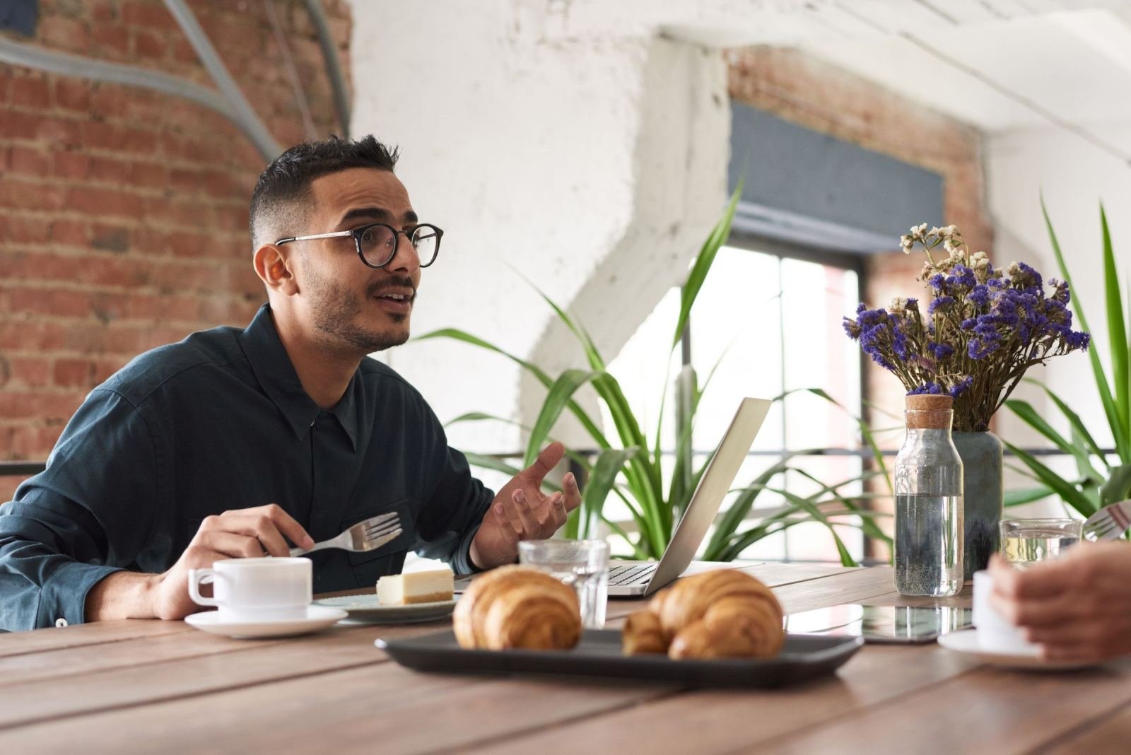 A man with glasses gestures while talking at a table with a laptop and breakfast