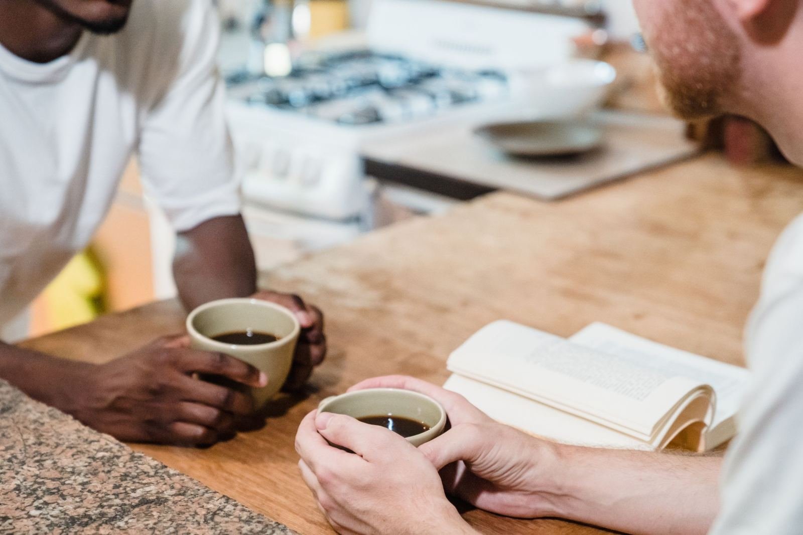 Two men hold cups of coffee while sitting at a wooden counter with an open notebook