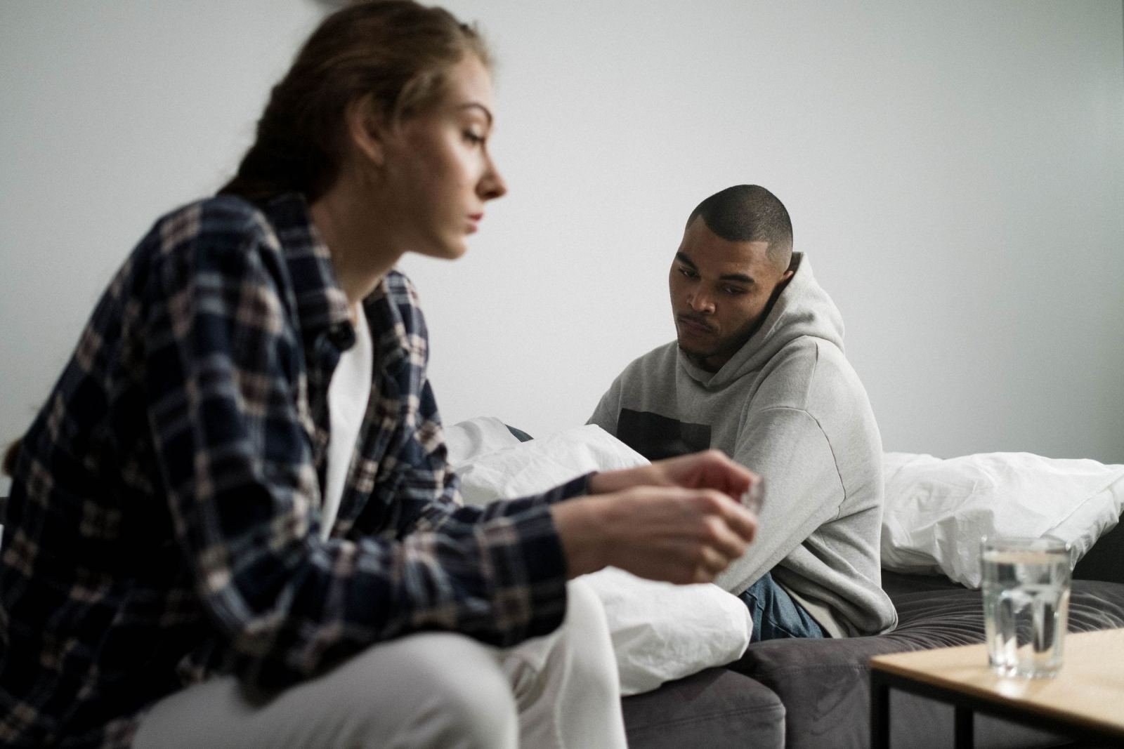 A man sits on a couch while a woman in a plaid shirt speaks with him.
