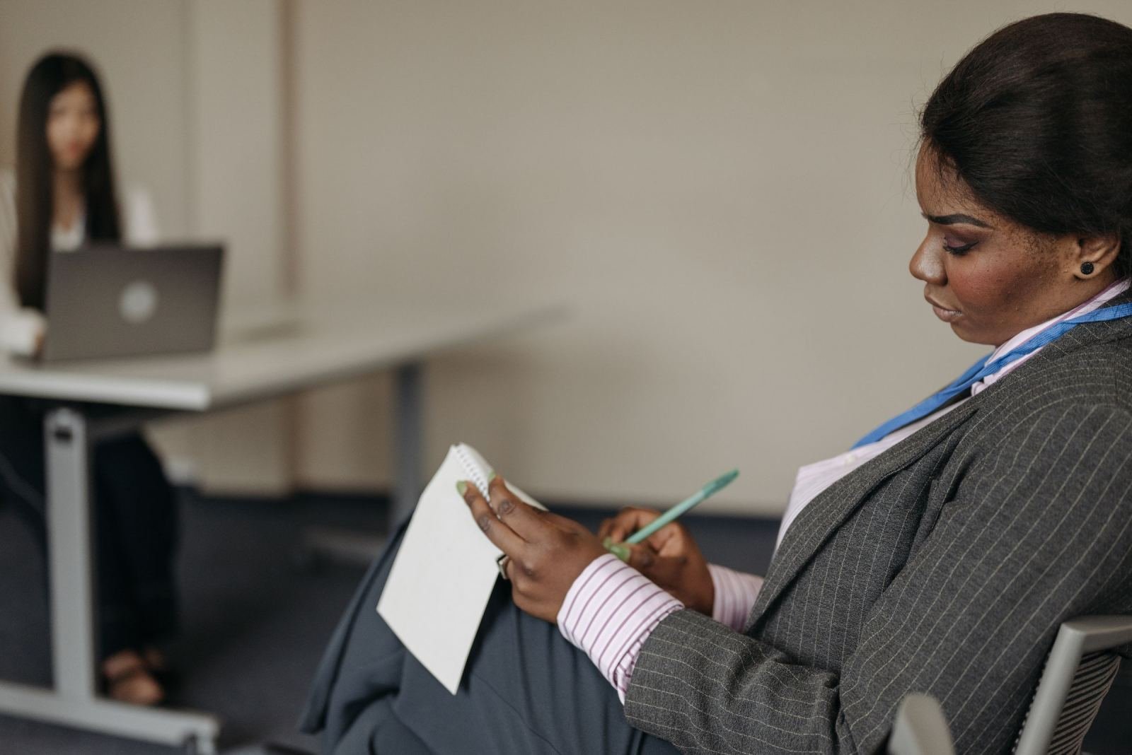 A woman in a blazer writes in a notebook while another person works at a desk in the background