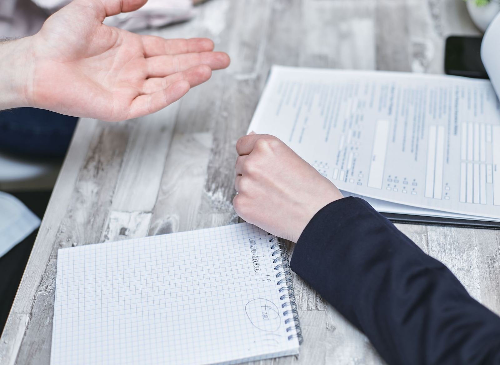 Two people sit at a wooden table with a notebook and a printed document