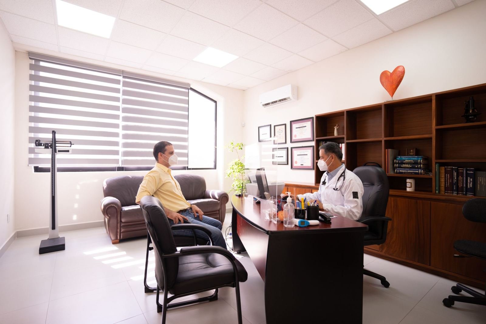 A clinician in a white coat and mask speaks with a patient in a bright office