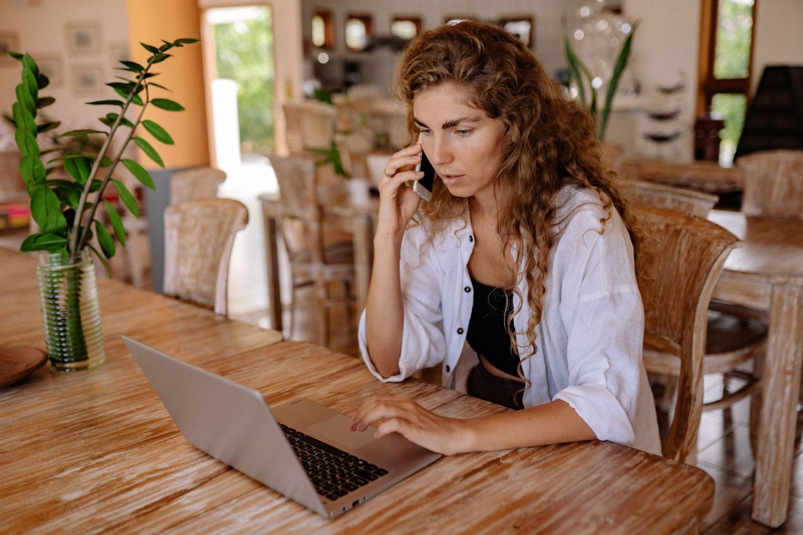 A woman on a phone call looks at a laptop while sitting at a wooden table