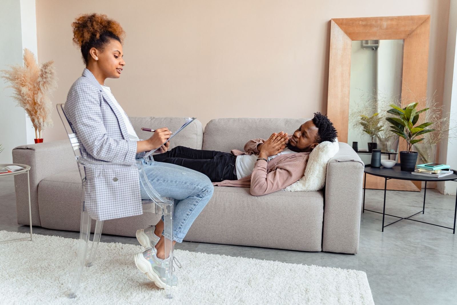 A man lies on a grey couch while a therapist holding a clipboard speaks with him.