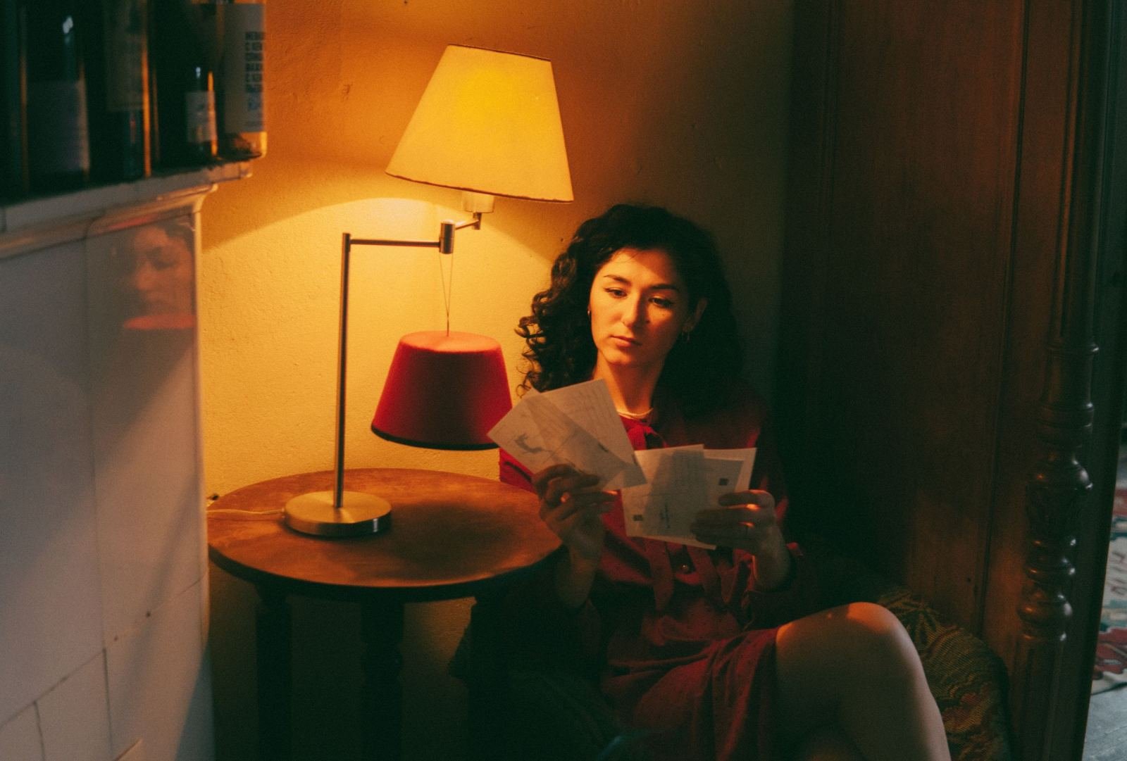 A woman sits in a dimly lit room reading papers by the light of a lamp