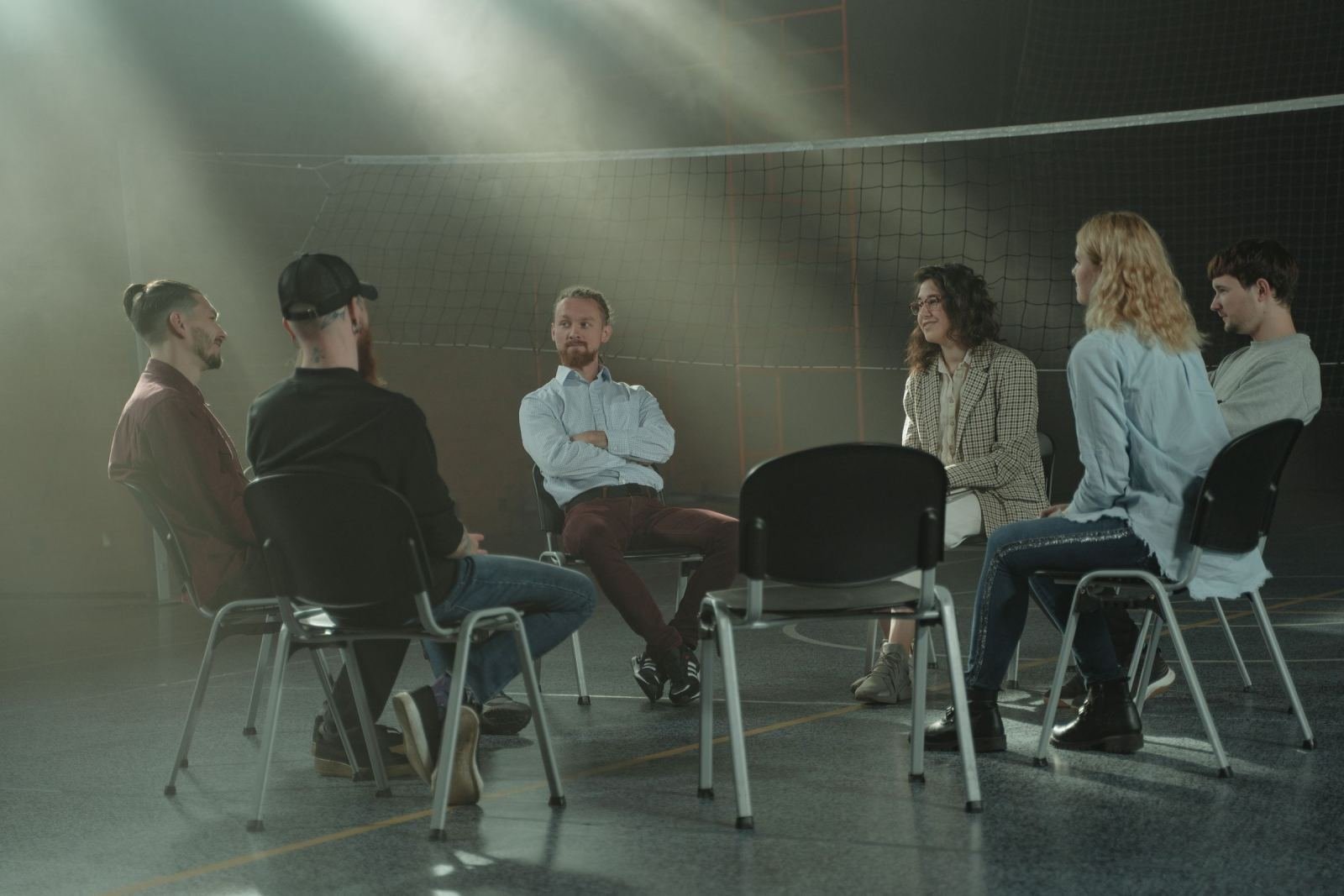 Five people sit in a circle of chairs in a dim gym with light beams shining down
