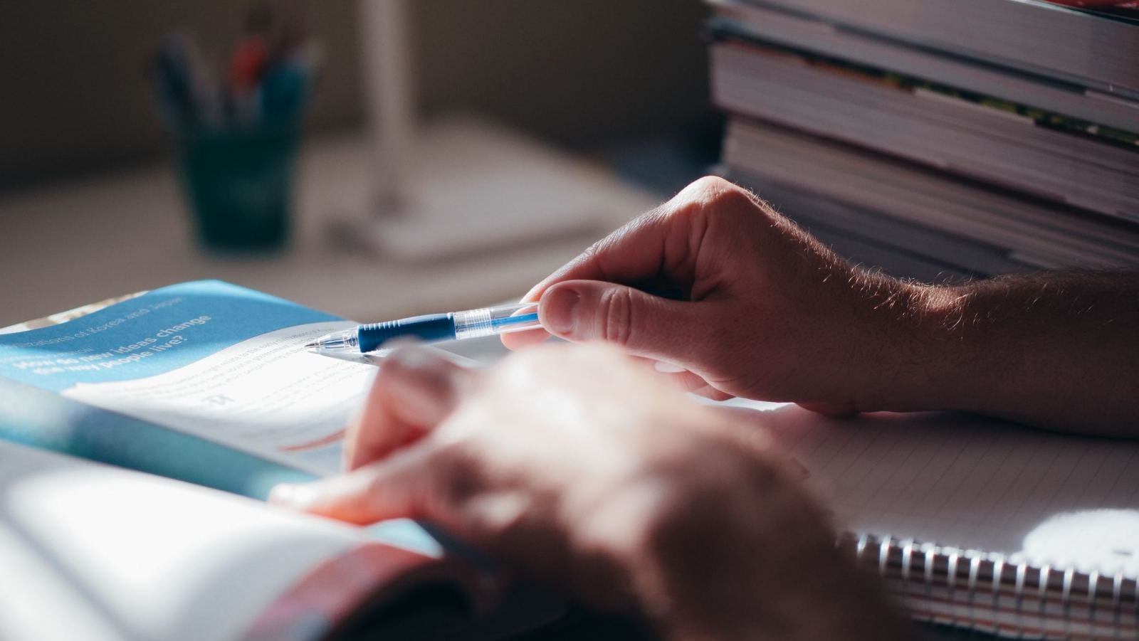 A person uses a blue pen to point to text in an open book on a desk