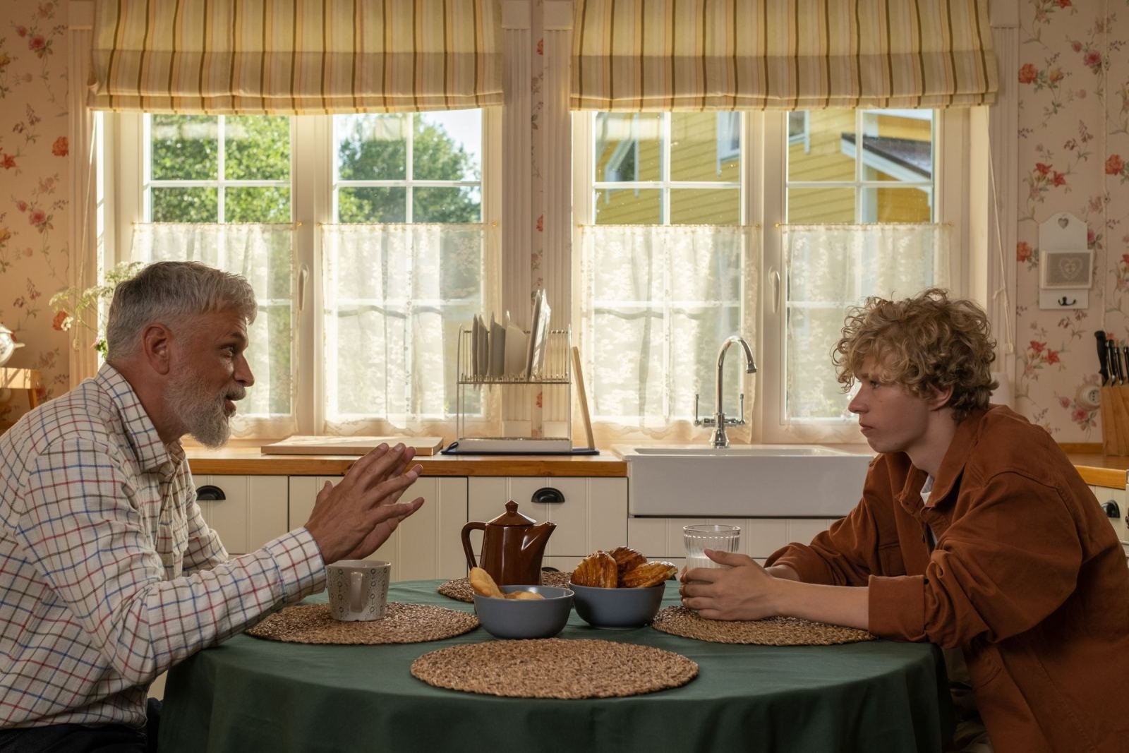 A man with a grey beard talks to a younger person across a kitchen table