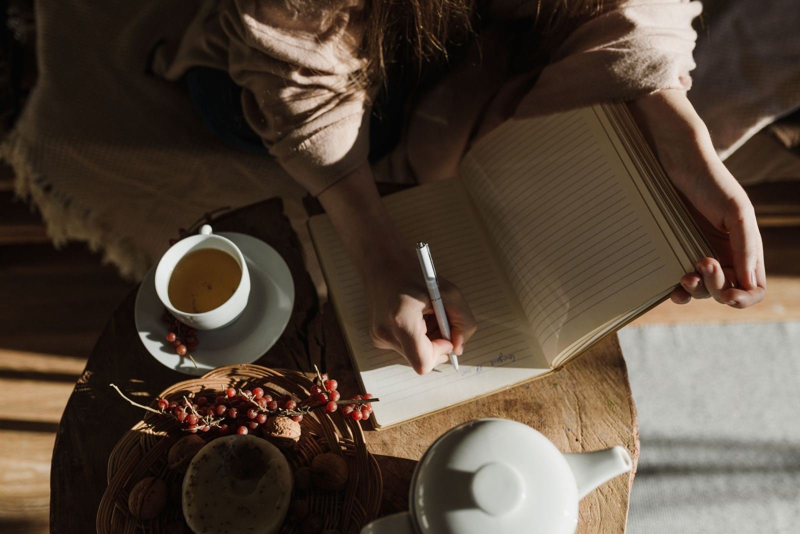 A person writing in a notebook next to a cup of tea and a teapot
