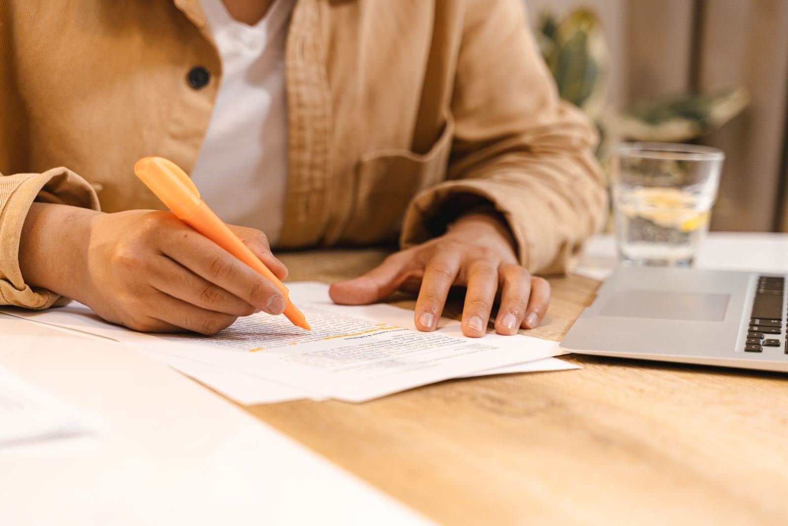 Someone marks a paper with an orange highlighter while sitting at a wood desk.