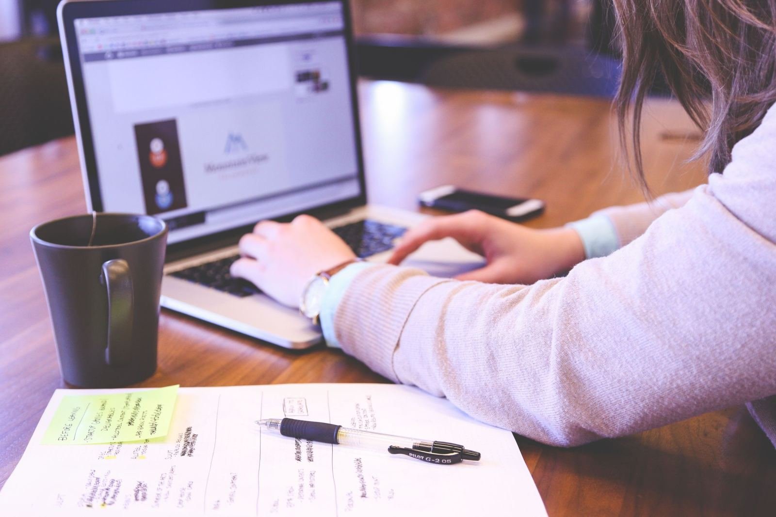 A person uses a laptop and writes notes in a notebook at a wooden desk